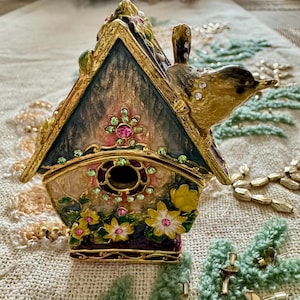 May include: A gold birdhouse with a bird perched on the roof. The birdhouse is decorated with flowers, leaves, and rhinestones. The birdhouse is a decorative piece and is not functional.