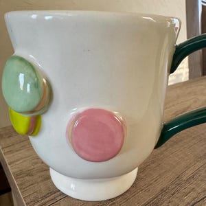 K&ouml;nnte beinhalten: Wei&szlig;e Keramiktasse mit gr&uuml;nem Henkel und dekorativen, farbenfrohen, runden Akzenten. Die Tasse hat einen breiten Boden und eine gl&auml;nzende Oberfl&auml;che. Die Akzente umfassen gr&uuml;ne, rosa und gelbe Kreise und verleihen dem Design eine verspielte Note.