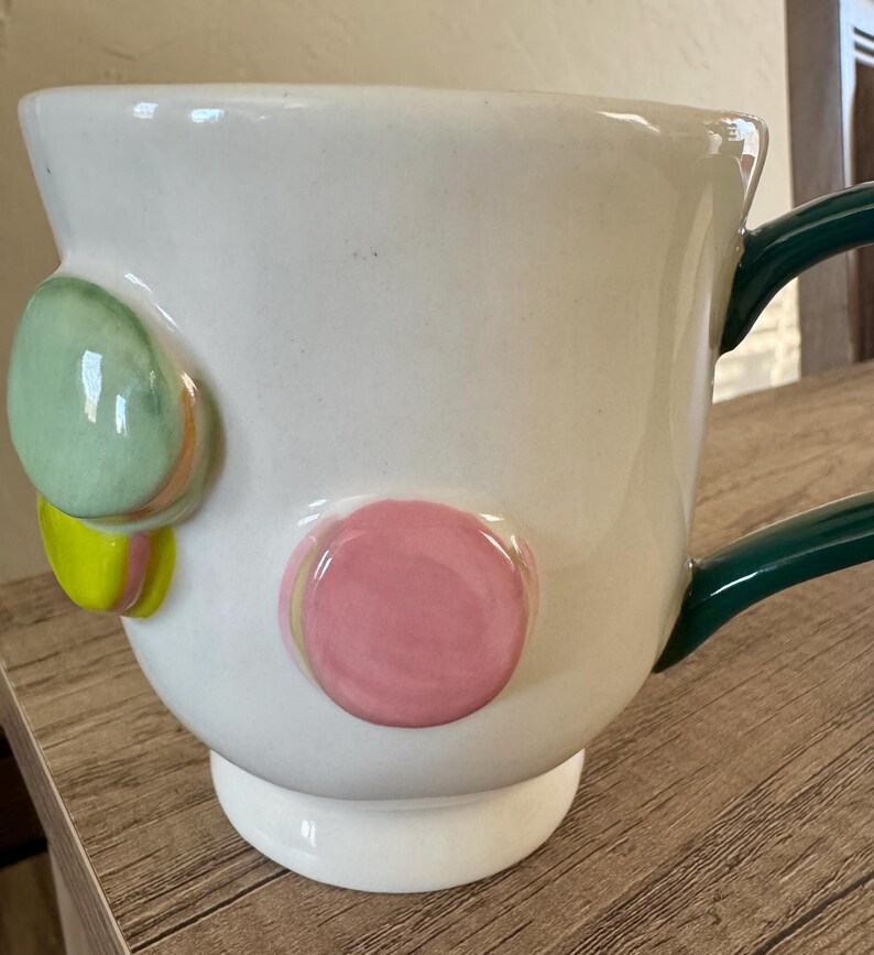 K&ouml;nnte beinhalten: Wei&szlig;e Keramiktasse mit gr&uuml;nem Henkel und dekorativen, farbenfrohen, runden Akzenten. Die Tasse hat einen breiten Boden und eine gl&auml;nzende Oberfl&auml;che. Die Akzente umfassen gr&uuml;ne, rosa und gelbe Kreise und verleihen dem Design eine verspielte Note.