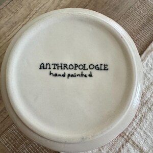May include: A white ceramic bowl with the text "ANTHROPOLOGIE hand painted" in black. The bowl has a slightly rounded shape and a red rim. The bowl is sitting on a white and beige surface.