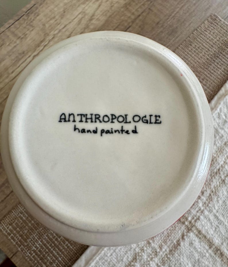 May include: A white ceramic bowl with the text "ANTHROPOLOGIE hand painted" in black. The bowl has a slightly rounded shape and a red rim. The bowl is sitting on a white and beige surface.
