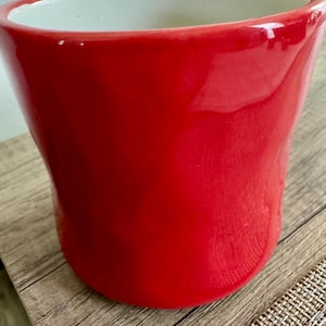 May include: A bright red ceramic plant pot with a glossy finish. The pot has a wide opening and a gently curved shape, with a white interior. Suitable for indoor or outdoor use, it is a decorative item.