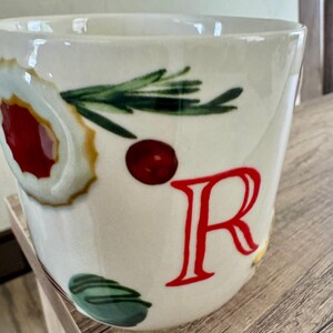 May include: White ceramic mug with a festive design. The mug features a large red letter "R", a sprig of green rosemary, a red berry, and a decorative cookie design. The mug is perfect for hot beverages.