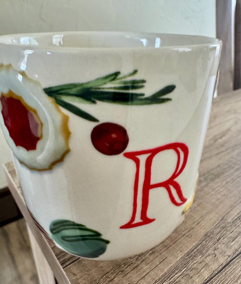 May include: White ceramic mug with a festive design. The mug features a large red letter "R", a sprig of green rosemary, a red berry, and a decorative cookie design. The mug is perfect for hot beverages.