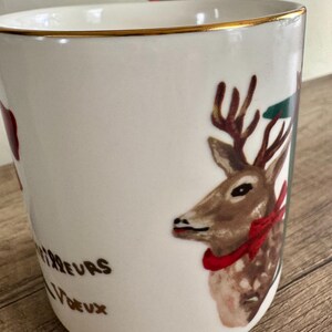 Puede incluir: Taza de cer&aacute;mica blanca con borde dorado, con una ilustraci&oacute;n de ciervo con lazo rojo y cinta verde y roja. La taza tiene el texto "Meilleurs Voeux" en letras doradas. Un dise&ntilde;o festivo navide&ntilde;o.
