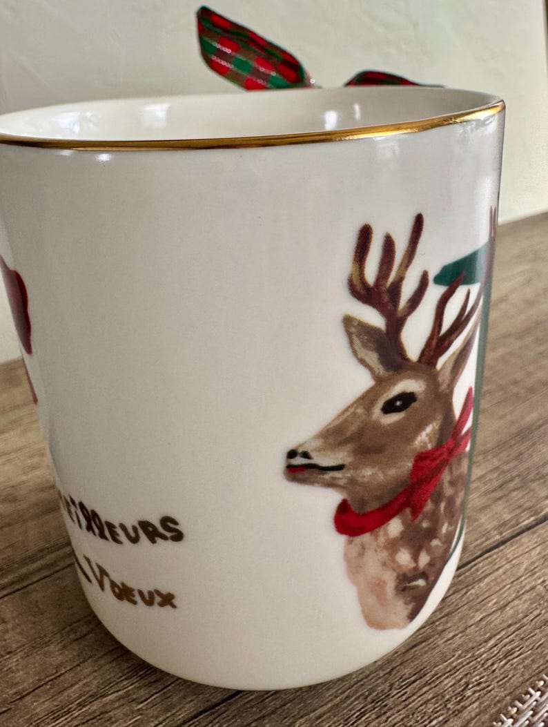 Puede incluir: Taza de cer&aacute;mica blanca con borde dorado, con una ilustraci&oacute;n de ciervo con lazo rojo y cinta verde y roja. La taza tiene el texto "Meilleurs Voeux" en letras doradas. Un dise&ntilde;o festivo navide&ntilde;o.