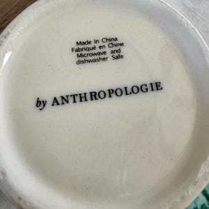 K&ouml;nnte beinhalten: Nahaufnahme des Bodens einer wei&szlig;en Keramiktasse. Der Text "Made in China, Fabriqu&eacute; en Chine, Microwave and dishwasher Safe" ist schwarz gedruckt. Die Worte "by ANTHROPOLOGIE" sind ebenfalls schwarz gedruckt.