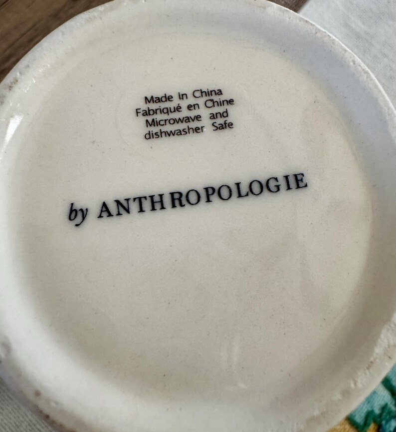 K&ouml;nnte beinhalten: Nahaufnahme des Bodens einer wei&szlig;en Keramiktasse. Der Text "Made in China, Fabriqu&eacute; en Chine, Microwave and dishwasher Safe" ist schwarz gedruckt. Die Worte "by ANTHROPOLOGIE" sind ebenfalls schwarz gedruckt.