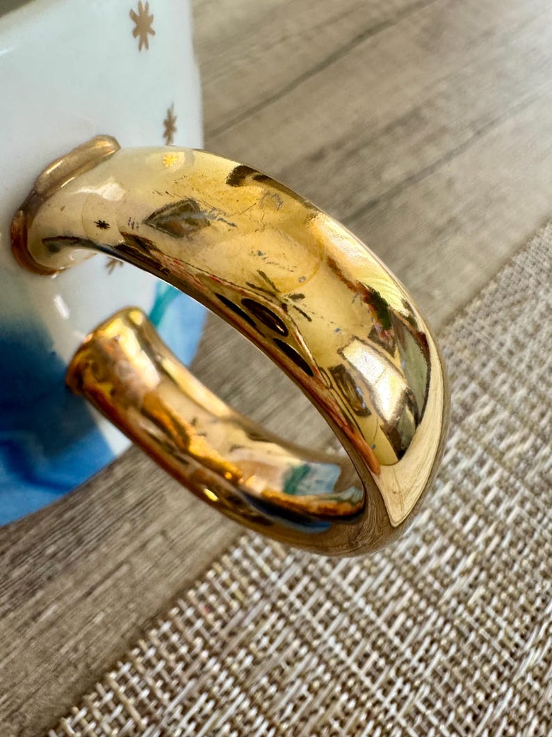 May include: A ceramic mug handle with a reflective gold metallic finish. The mug body features a white surface decorated with small gold star patterns and a blue gradient design visible near the base of the handle attachment points.
