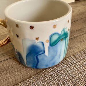 May include: A ceramic mug with a white interior and a glossy finish. The exterior features a watercolor-style pattern of blue and teal drips against a white background, accented by small, scattered gold star-shaped icons and a gold-colored handle.
