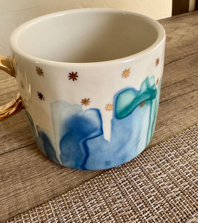 May include: A ceramic mug with a white interior and a glossy finish. The exterior features a watercolor-style pattern of blue and teal drips against a white background, accented by small, scattered gold star-shaped icons and a gold-colored handle.