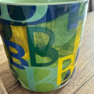 May include: A green ceramic planter with a colorful abstract design featuring letters and shapes in shades of blue, yellow, and green.