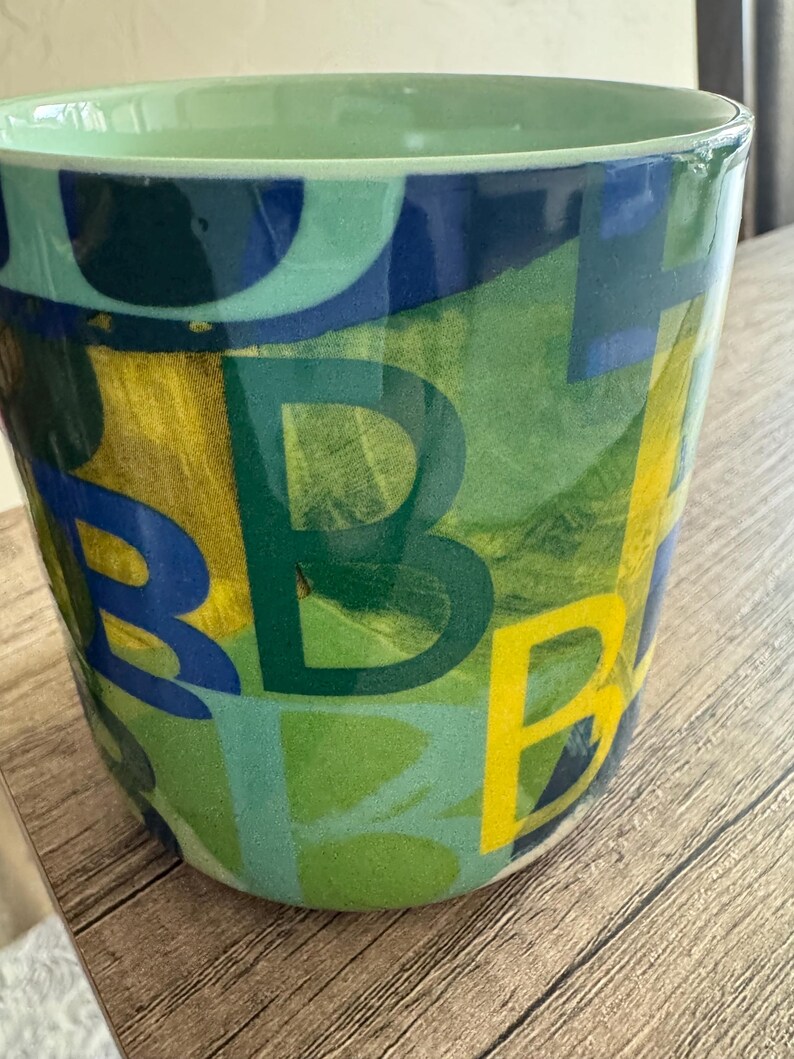 May include: A green ceramic planter with a colorful abstract design featuring letters and shapes in shades of blue, yellow, and green.