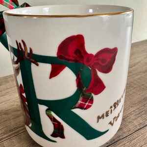 Puede incluir: Taza de cer&aacute;mica blanca con borde dorado, con una gran letra "R" verde adornada con un lazo rojo y detalles de cuadros escoceses. La taza tiene un asa verde y una cinta decorativa. Las palabras "Merry Christmas" est&aacute;n escritas en el lateral.