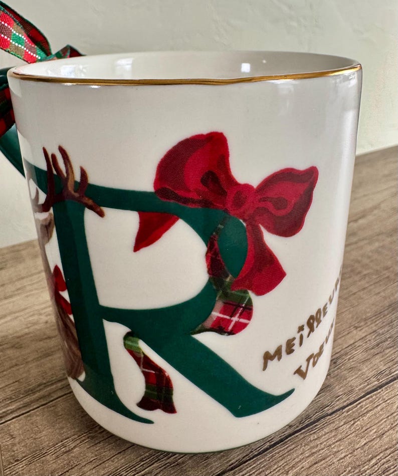 Puede incluir: Taza de cer&aacute;mica blanca con borde dorado, con una gran letra "R" verde adornada con un lazo rojo y detalles de cuadros escoceses. La taza tiene un asa verde y una cinta decorativa. Las palabras "Merry Christmas" est&aacute;n escritas en el lateral.