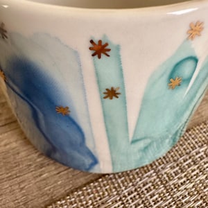 May include: A white ceramic mug featuring a watercolor-style design with blended shades of blue and teal. The surface is decorated with small, metallic gold star-shaped accents scattered across the painted blue and teal brushstroke patterns.