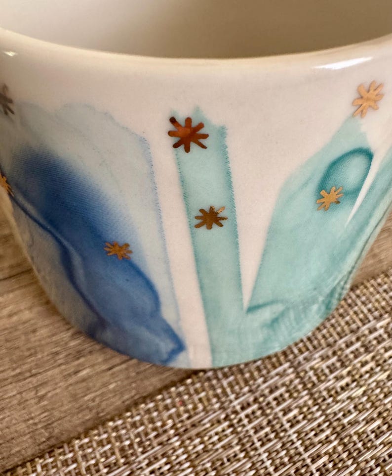May include: A white ceramic mug featuring a watercolor-style design with blended shades of blue and teal. The surface is decorated with small, metallic gold star-shaped accents scattered across the painted blue and teal brushstroke patterns.