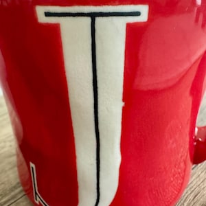 May include: A red ceramic mug featuring a large white letter "J" in the centre, outlined in black. The mug has a glossy finish and a handle on the right side.