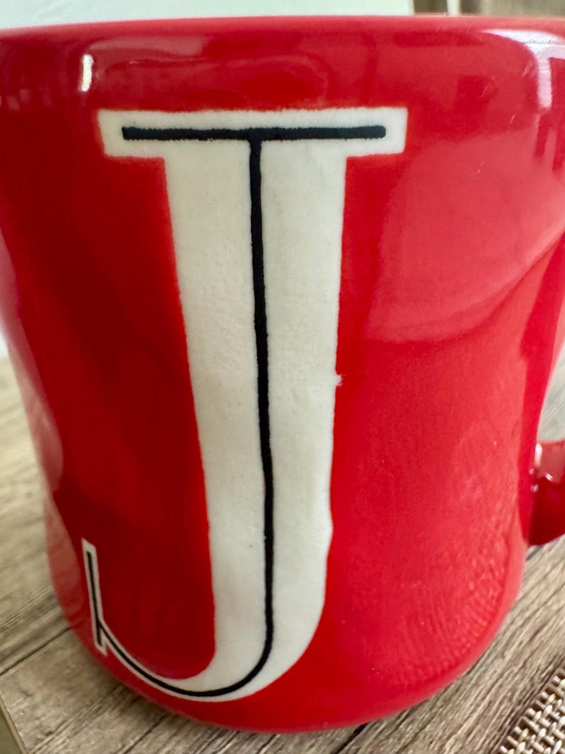 May include: A red ceramic mug featuring a large white letter "J" in the centre, outlined in black. The mug has a glossy finish and a handle on the right side.