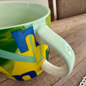 May include: A ceramic mug with a light green interior and handle. The exterior features a vibrant design with yellow, blue, and green abstract shapes. The mug is set on a wooden surface.