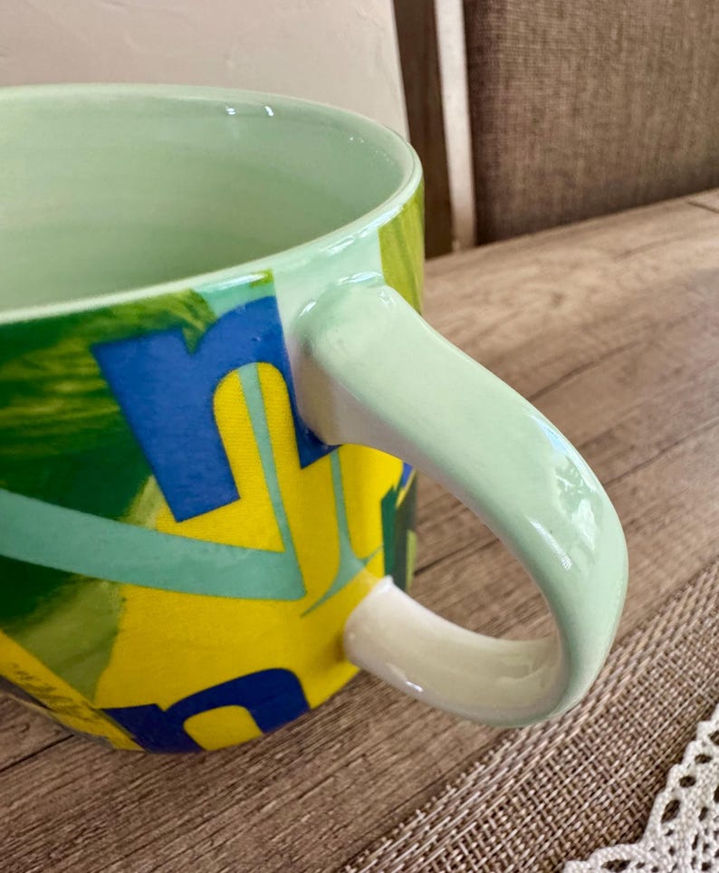 May include: A ceramic mug with a light green interior and handle. The exterior features a vibrant design with yellow, blue, and green abstract shapes. The mug is set on a wooden surface.