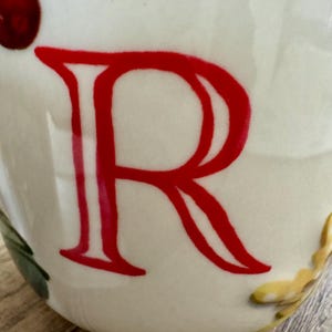 May include: A white ceramic mug featuring a large red letter "R". The mug is decorated with red berries and green leaves. The mug has a rounded shape. The mug is on a wooden surface.