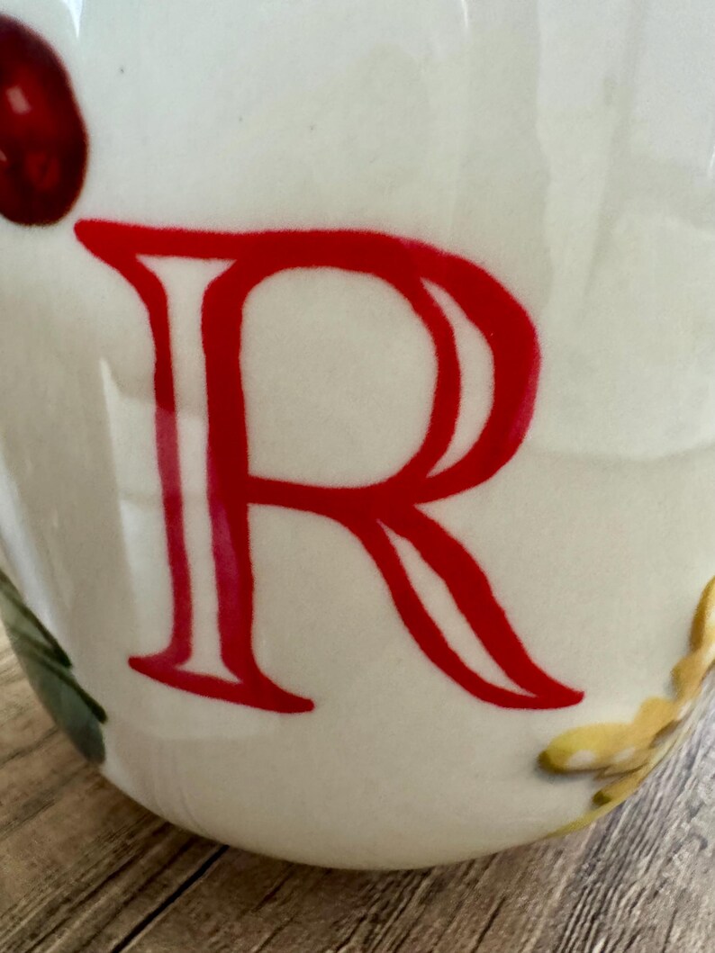 May include: A white ceramic mug featuring a large red letter "R". The mug is decorated with red berries and green leaves. The mug has a rounded shape. The mug is on a wooden surface.