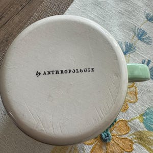 May include: White ceramic mug with a green handle. The mug has the text "by Anthropologie" printed on the bottom.