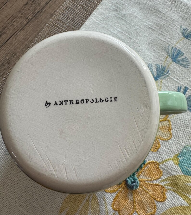 May include: White ceramic mug with a green handle. The mug has the text "by Anthropologie" printed on the bottom.