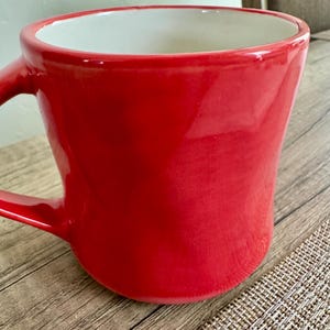 May include: A vibrant red ceramic mug with a white interior. The mug has a curved shape and a sturdy handle. The glossy finish reflects light, highlighting the smooth surface. This mug is perfect for enjoying your favorite hot beverages.
