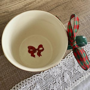 Puede incluir: Cuenco de cer&aacute;mica color crema con borde dorado y dise&ntilde;o de lazo rojo en el interior. Un soporte de cer&aacute;mica verde y una cinta a cuadros roja, verde y dorada est&aacute;n adjuntos, sugiriendo un tema festivo.