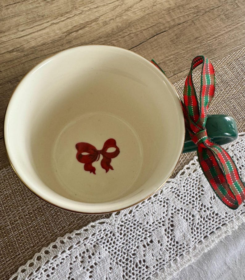 Puede incluir: Cuenco de cer&aacute;mica color crema con borde dorado y dise&ntilde;o de lazo rojo en el interior. Un soporte de cer&aacute;mica verde y una cinta a cuadros roja, verde y dorada est&aacute;n adjuntos, sugiriendo un tema festivo.