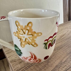 May include: White ceramic mug with festive holiday designs. The mug features a large yellow snowflake, red and green swirls, and other colourful accents. The handle has a red and white pattern.