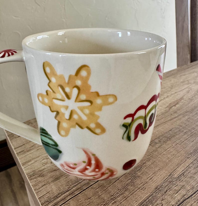 May include: White ceramic mug with festive holiday designs. The mug features a large yellow snowflake, red and green swirls, and other colourful accents. The handle has a red and white pattern.