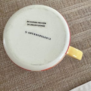 May include: A white ceramic mug with a red and yellow rim and handle. The bottom of the mug has the text "by ANTHROPOLOGIE" and "DISHWASHER & MICROWAVE SAFE MADE IN CHINA / FABRIQUE EN CHINE".