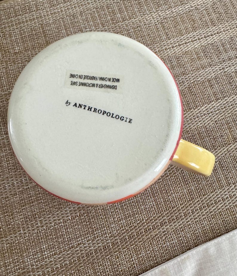 May include: A white ceramic mug with a red and yellow rim and handle. The bottom of the mug has the text "by ANTHROPOLOGIE" and "DISHWASHER & MICROWAVE SAFE MADE IN CHINA / FABRIQUE EN CHINE".