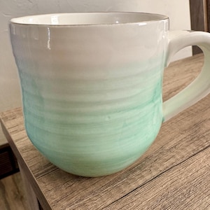 Pode incluir: Uma caneca de cer&acirc;mica com um topo branco que se desvanece em uma cor verde-&aacute;gua. A caneca tem uma forma arredondada e uma al&ccedil;a grande. A caneca est&aacute; em uma superf&iacute;cie de madeira.