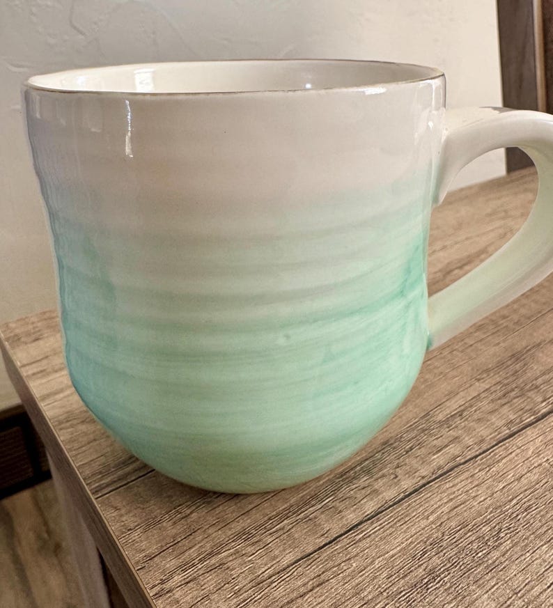 Pode incluir: Uma caneca de cer&acirc;mica com um topo branco que se desvanece em uma cor verde-&aacute;gua. A caneca tem uma forma arredondada e uma al&ccedil;a grande. A caneca est&aacute; em uma superf&iacute;cie de madeira.