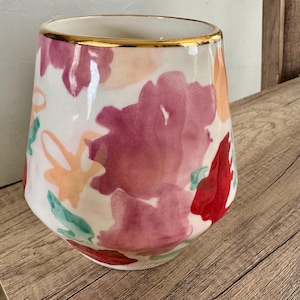 May include: A stemless wine glass with a white base, decorated with watercolour floral designs in red, pink, and peach hues. The rim is trimmed with a gold band. The glass sits on a wooden surface.