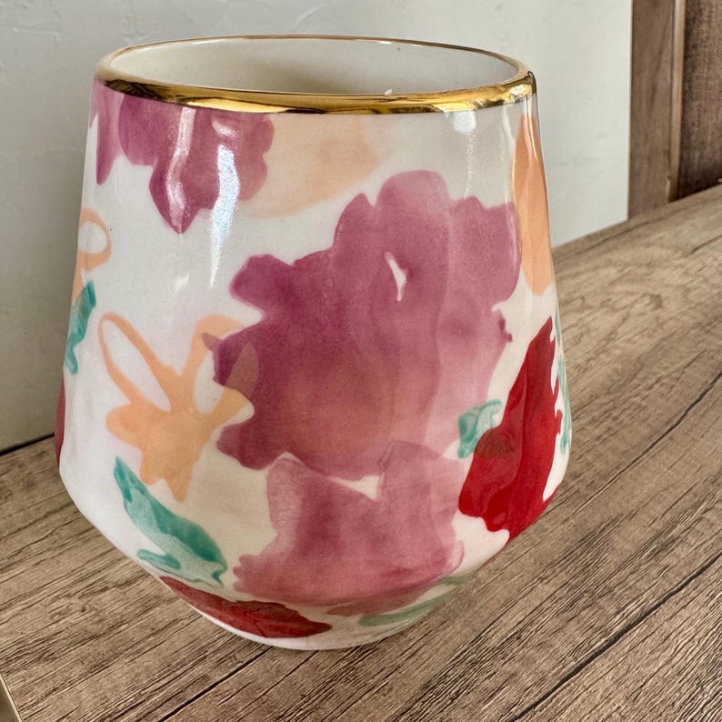 May include: A stemless wine glass with a white base, decorated with watercolour floral designs in red, pink, and peach hues. The rim is trimmed with a gold band. The glass sits on a wooden surface.