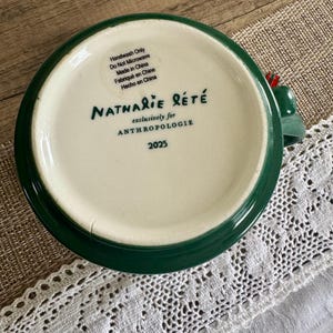Puede incluir: Una taza de cer&aacute;mica verde y blanca con el texto "Nathalie Let&eacute;" y "exclusively for Anthropologie" impreso en la parte inferior. La taza tambi&eacute;n tiene el n&uacute;mero "2025" impreso. La taza est&aacute; etiquetada como "Handwash Only" y "Do Not Microwave".