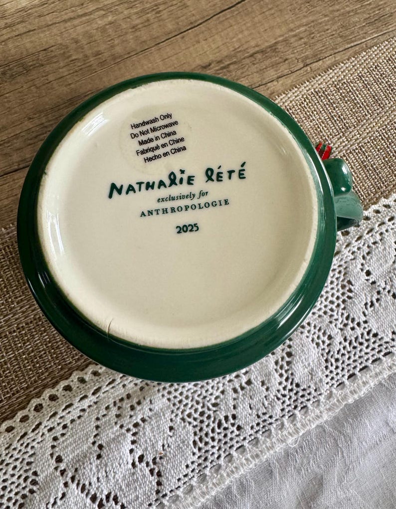 Puede incluir: Una taza de cer&aacute;mica verde y blanca con el texto "Nathalie Let&eacute;" y "exclusively for Anthropologie" impreso en la parte inferior. La taza tambi&eacute;n tiene el n&uacute;mero "2025" impreso. La taza est&aacute; etiquetada como "Handwash Only" y "Do Not Microwave".