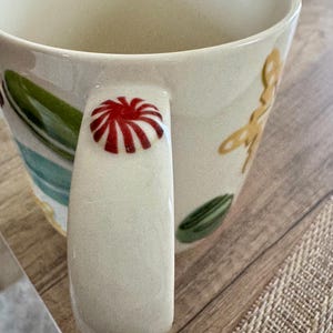 May include: Cream ceramic mug with a red and white design on the handle. The mug features green and yellow floral and leaf patterns. The mug is on a wooden surface.
