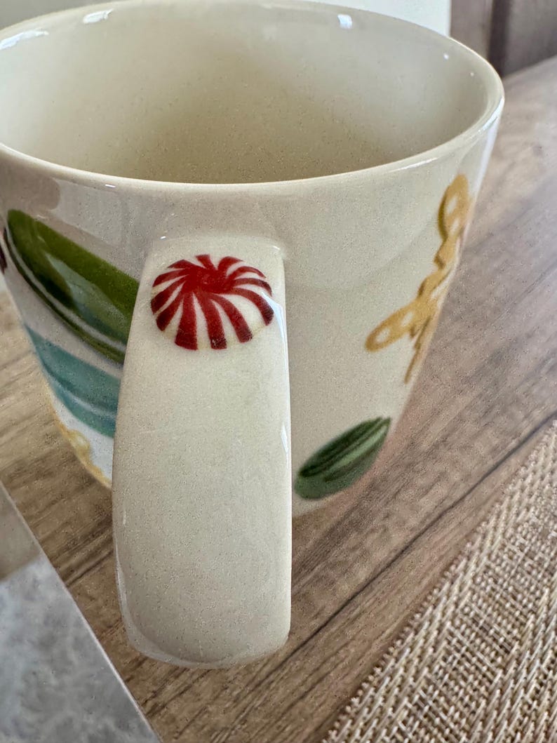 May include: Cream ceramic mug with a red and white design on the handle. The mug features green and yellow floral and leaf patterns. The mug is on a wooden surface.