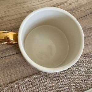 May include: A white ceramic mug featuring a smooth, glossy interior and a reflective metallic gold handle. The mug is viewed from directly above, showcasing its circular rim, deep basin, and the curved shape of the gold-toned handle extending outward.