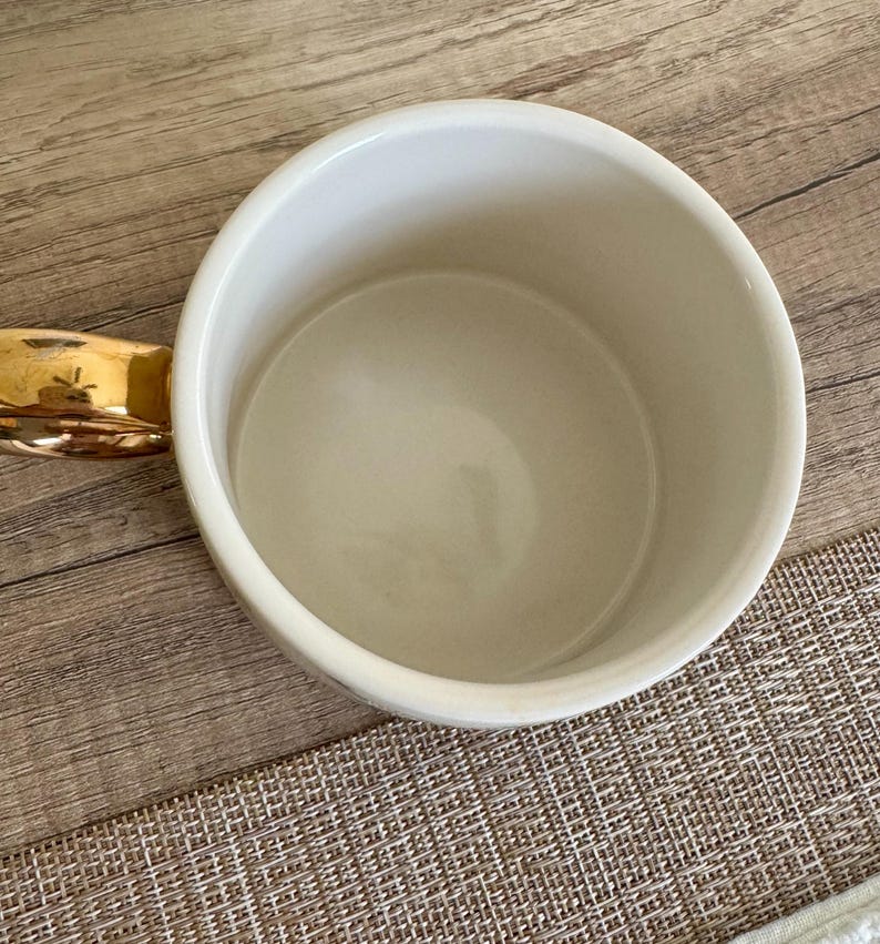 May include: A white ceramic mug featuring a smooth, glossy interior and a reflective metallic gold handle. The mug is viewed from directly above, showcasing its circular rim, deep basin, and the curved shape of the gold-toned handle extending outward.