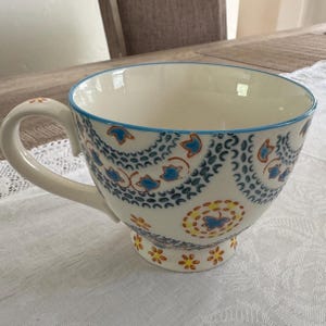 May include: A large, white ceramic teacup with a blue rim and handle. The cup features a detailed pattern of blue and orange floral designs. The base of the cup has a small pedestal with matching floral accents.