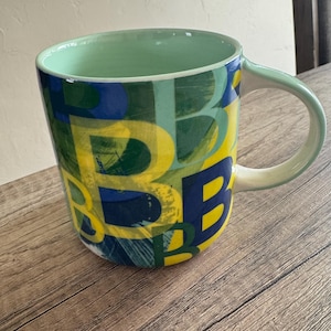 May include: A large ceramic mug with a green rim and handle. The mug is decorated with a colorful abstract pattern of yellow, blue, and green letters.