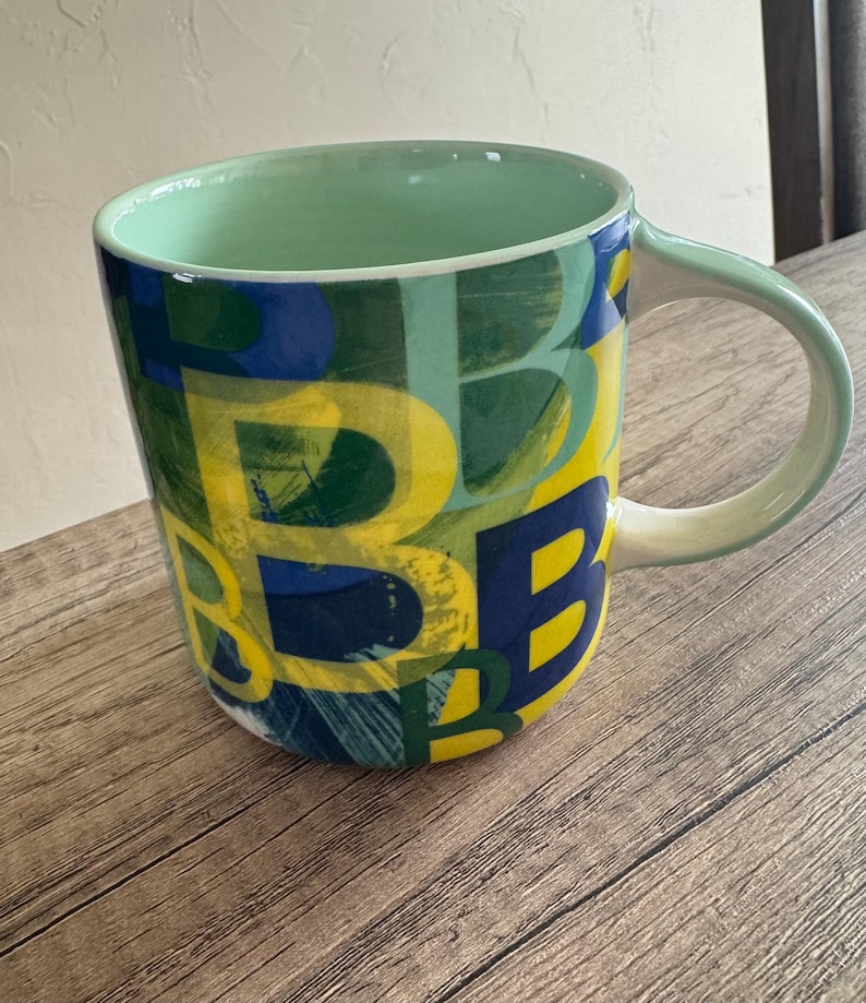 May include: A large ceramic mug with a green rim and handle. The mug is decorated with a colorful abstract pattern of yellow, blue, and green letters.