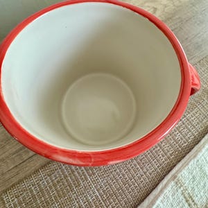 May include: A ceramic mug with a wide opening and a red rim. The interior is white, and the mug has a handle on the side. The mug is sitting on a textured surface.